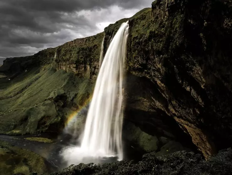 A majestic waterfall cascades over a rocky cliff, creating a misty rainbow in the lush, green landscape.