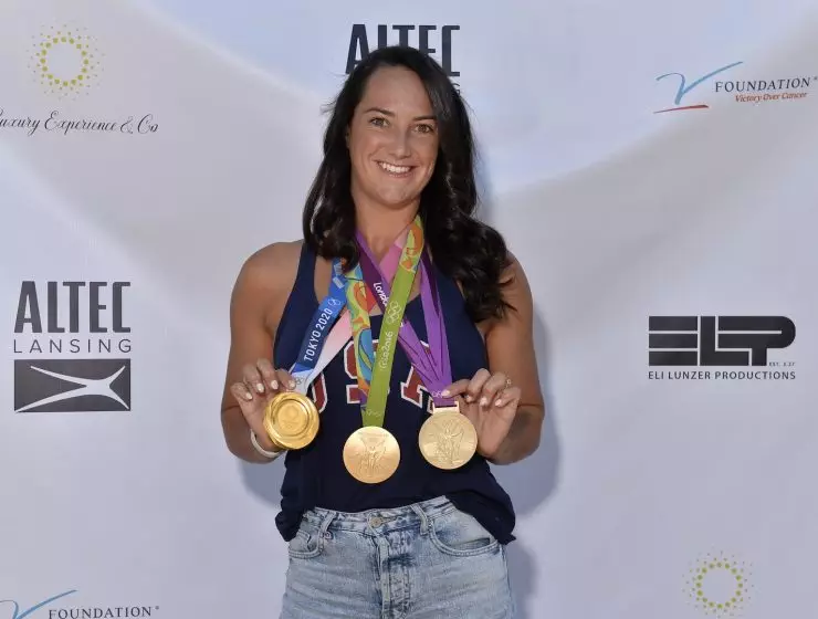 A smiling woman displaying multiple medals around her neck at an outdoor event with sponsor banners in the background.