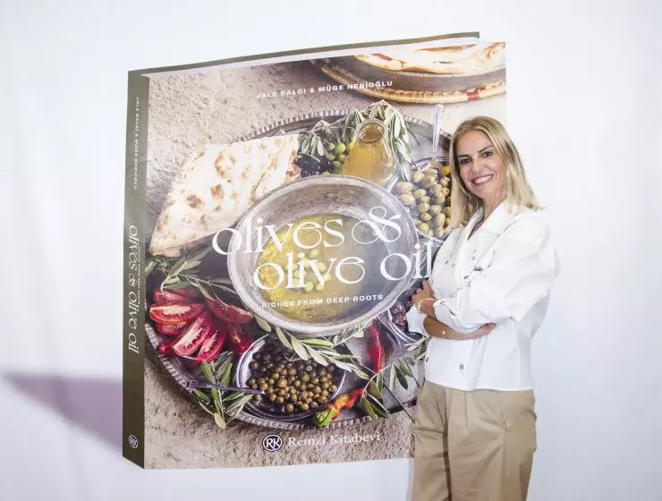 A woman is standing next to a large displayed book titled Olives & Olive Oil with images of olives, oil, and Mediterranean food.