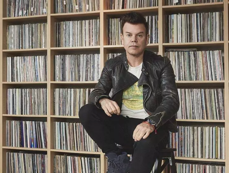 A man is sitting on a stool in front of a large bookshelf filled with records, wearing a black leather jacket and jeans.