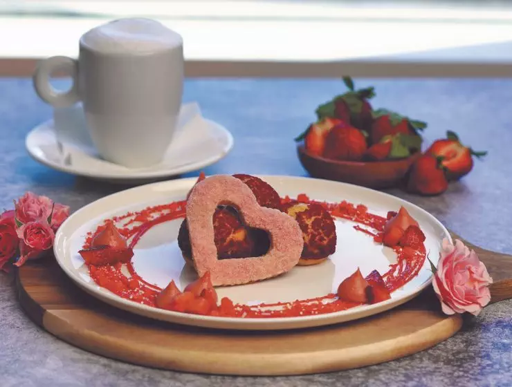 A romantic breakfast setup featuring a heart-shaped egg, pastries, strawberries, flowers, and a cup of cappuccino on a wooden tray.