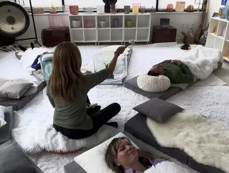 A group of people is lying on mats in a room with large windows offering a mountain view, participating in a relaxation or meditation session.