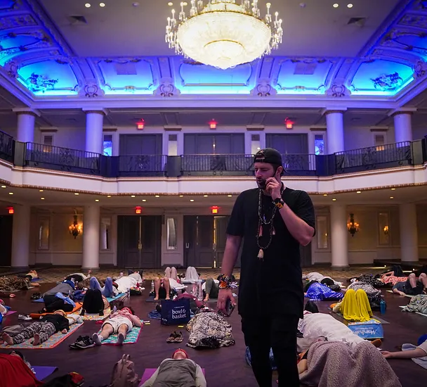 A man stands on a stage speaking on a phone, overlooking a room of people lying on the floor in a relaxed pose under a large chandelier.