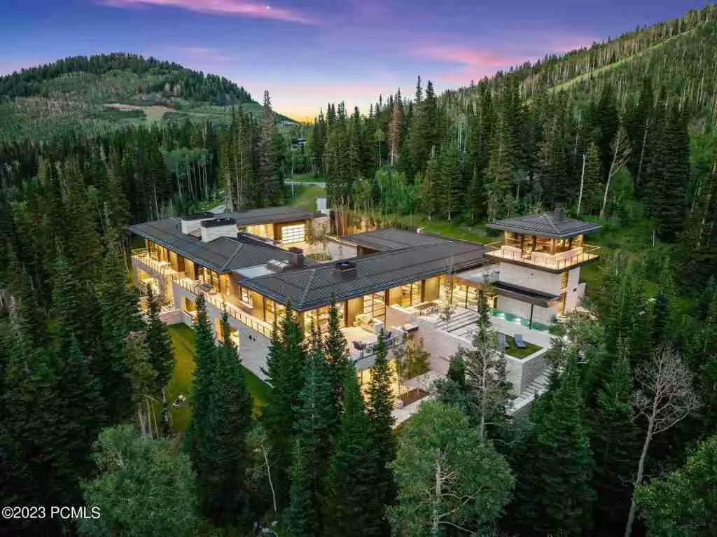 A luxurious modern house with bright interior lights surrounded by dense forest under an evening sky.